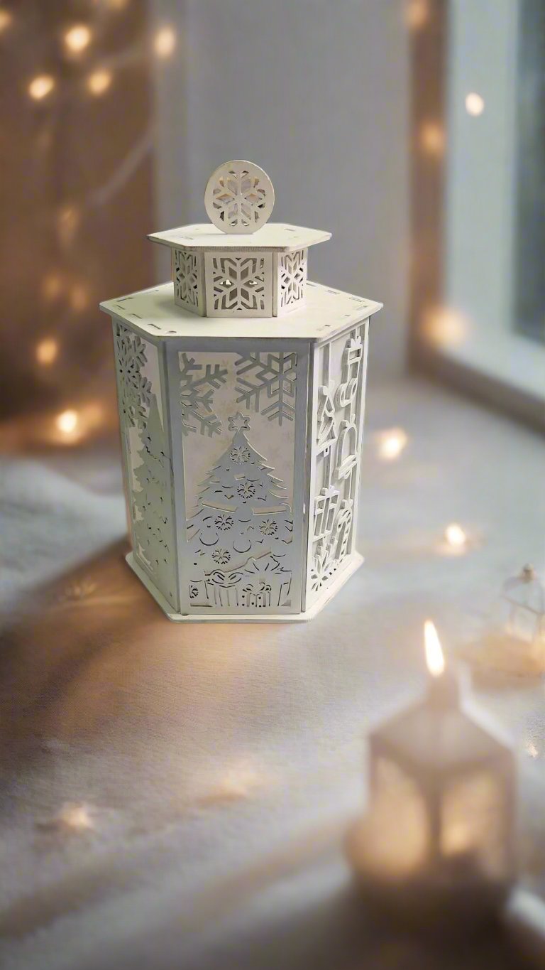 A white tea light candle holder with a Christmas tree and snowflake design, placed on a surface with a blurred Christmas tree in the background.