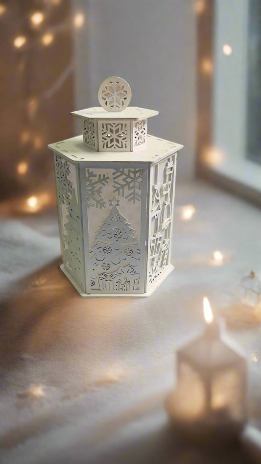 A white tea light candle holder with a Christmas tree and snowflake design, placed on a surface with a blurred Christmas tree in the background.