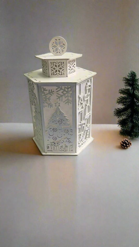A white tea light candle holder with a Christmas tree and snowflake design, placed on a surface with a blurred Christmas tree in the background.