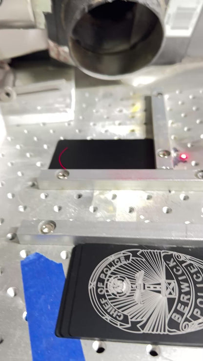 a video showing the making of A black metal business card with white and pink text, displaying the name of the engraving service and contact information.